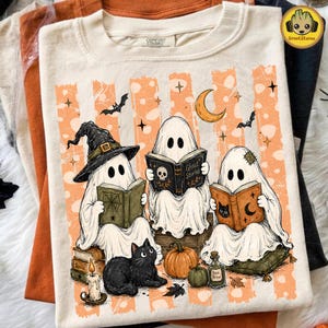 May include: A cream-colored t-shirt featuring a graphic of three white ghosts reading books. The ghosts are surrounded by orange vertical stripes, a crescent moon, bats, a black cat, a pumpkin, and a candle. The center book is titled Ghost Stories.