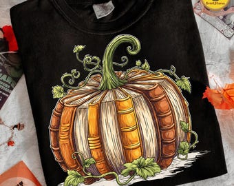 May include: Black t-shirt featuring a detailed illustration of a pumpkin made of books. The pumpkin has a green stem and vines with leaves. The books are brown and beige, creating a unique Halloween or fall-themed design.