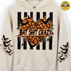May include: A beige hooded sweatshirt featuring a central graphic of an orange heart filled with spiderwebs and black bats. The text BAT SHIT CRAZY is printed across the center over black vertical stripes. Both sleeves feature black bat silhouettes.