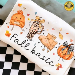 May include: A white t-shirt featuring a colourful graphic print of a smiling orange beanie with the word FALL, a leopard print tumbler with a bow, tan boots, and an orange pumpkin with a black and white plaid bow. The text Fall basic is printed below.