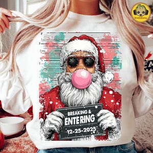 以下が含まれることがあります： サングラスをかけ、ガムを膨らませているサンタクロースのユーモラスなグラフィックが特徴の白いスウェットシャツ。「Breaking & Entering 12-25-2025」と書かれた看板を持っています。お祭り気分で面白いテーマです。