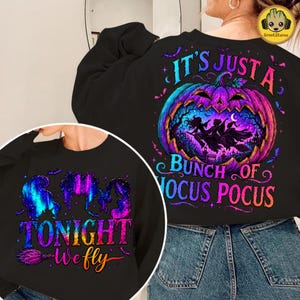 May include: A black long-sleeve sweatshirt featuring a large graphic of a purple and blue pumpkin with silhouettes of three figures flying on broomsticks inside. The text reads IT'S JUST A BUNCH OF HOCUS POCUS and TONIGHT WE FLY in colorful lettering.