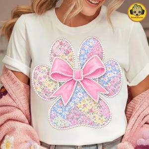 May include: White t-shirt featuring a floral paw print design with a pink bow. The paw print is made of pastel floral patterns in pink, blue, and yellow. The bow is a bright pink color with a pearl-like trim.
