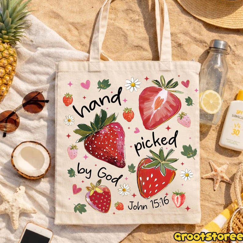 May include: A beige tote bag with a strawberry design and the text "hand picked by God John 15:16". The bag features illustrations of strawberries, leaves, and flowers. The bag has two handles.