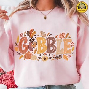 May include: A light pink crewneck sweatshirt featuring a graphic print with the text GOBBLE in large, stylized, multi-colored letters. The design includes a turkey head, pumpkins, autumn leaves, acorns, and a sunflower in shades of orange and brown.
