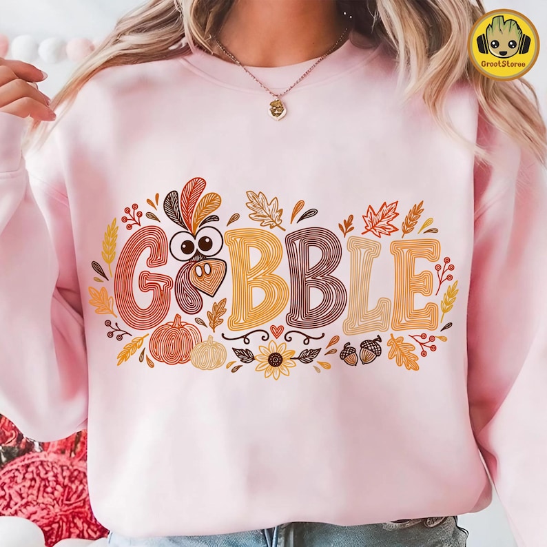 May include: A light pink crewneck sweatshirt featuring a graphic print with the text GOBBLE in large, stylized, multi-colored letters. The design includes a turkey head, pumpkins, autumn leaves, acorns, and a sunflower in shades of orange and brown.