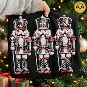 May include: A black sweatshirt featuring a graphic print of three nutcracker figures arranged in a row. Each nutcracker wears a tall black hat with red accents, a black uniform with red trim, and holds a staff, all rendered in a detailed illustration.