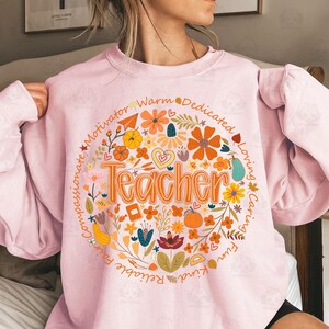 May include: A light pink sweatshirt featuring a floral design with the word "Teacher" in orange. The design includes flowers, hearts, and the words "Motivator", "Warm", "Dedicated", "Loving", "Caring", "Fun", "Kind", "Reliable", "Fair", and "Compassionate".