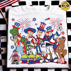 May include: A white t-shirt featuring a graphic print of Toy Story characters including Woody, Buzz Lightyear, Jessie, Rex, and others wearing patriotic red, white, and blue hats and accessories, surrounded by firework illustrations.