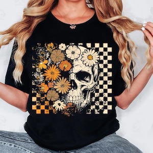 May include: Black t-shirt featuring a skull design with floral and checkered elements. The skull is partially obscured by orange, yellow, and white flowers. A checkered pattern is on the right side of the skull.