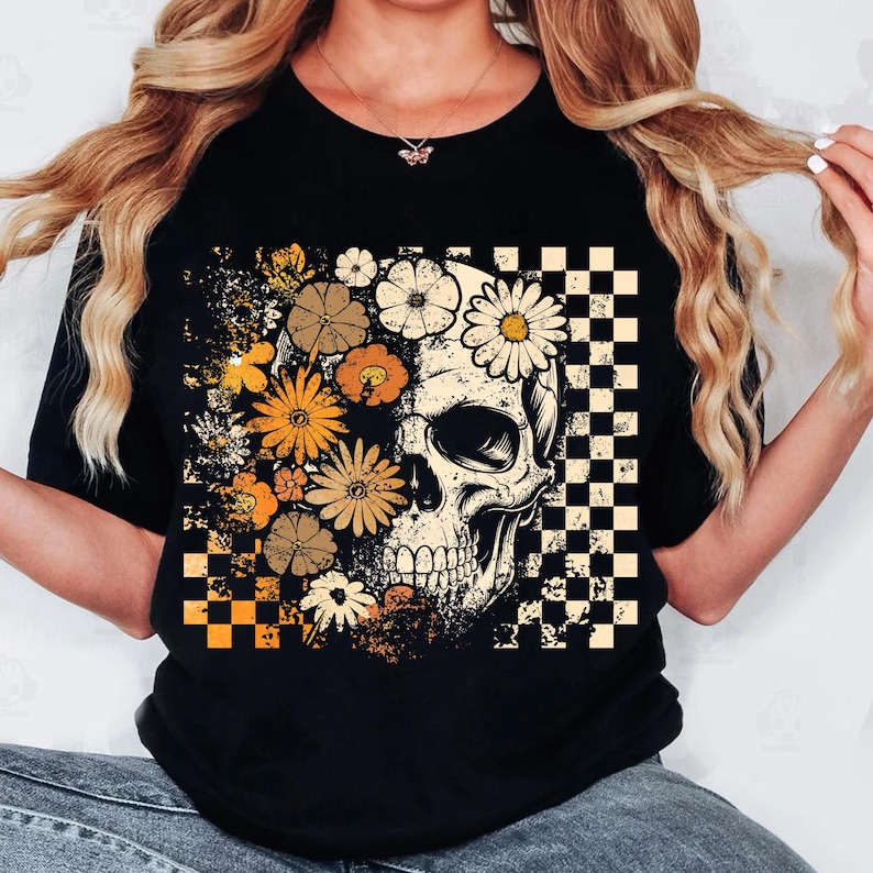 May include: Black t-shirt featuring a skull design with floral and checkered elements. The skull is partially obscured by orange, yellow, and white flowers. A checkered pattern is on the right side of the skull.