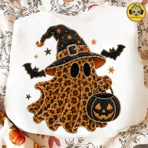 May include: A white sweatshirt featuring a graphic of a ghost wearing a black witch hat with orange stars. The ghost has an orange and black leopard print pattern and holds a black jack-o'-lantern bucket. Two black bats and orange stars surround the ghost.
