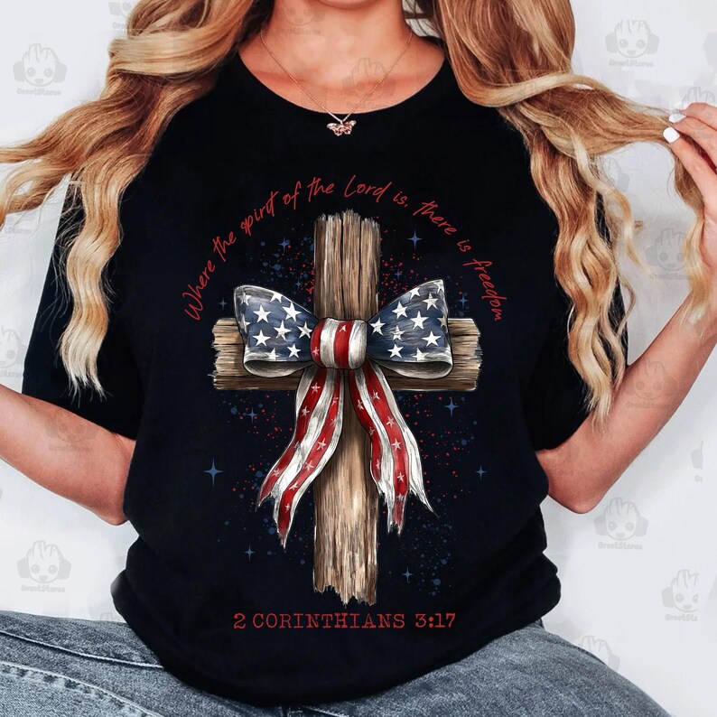 Może przedstawiać: Czarny t-shirt z drewnianym krzyżem ozdobionym kokardą w stylu amerykańskiej flagi. Koszulka zawiera napis "Where the spirit of the Lord is, there is freedom" i "2 Koryntian 3:17" w kolorze czerwonym.