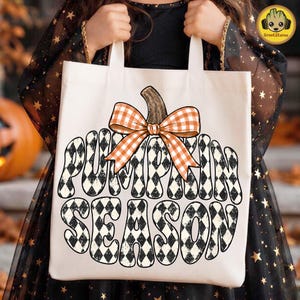 May include: A cream-colored canvas tote bag featuring a large graphic of a pumpkin shape filled with a black and white diamond pattern. The text PUMPKIN SEASON is written in a bold, wavy font within the pumpkin, topped with an orange gingham bow.