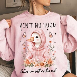 May include: Pink sweatshirt with a graphic of a duck wearing a pink hoodie and the text "Ain't no hood like motherhood" with flowers surrounding the duck.