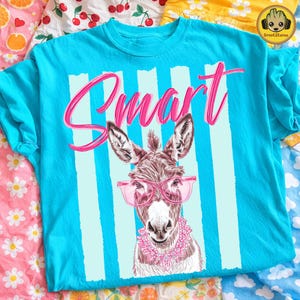 May include: A turquoise t-shirt with a graphic of a donkey wearing pink glasses and a floral collar. The word "Smart" is written in pink script. Light blue vertical stripes are behind the donkey.
