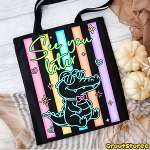 May include: Black tote bag with a colorful design featuring a cartoon alligator wearing sunglasses and a bow tie. The text "See you later" is written in a neon green font, with pink, blue, orange, and purple stripes.