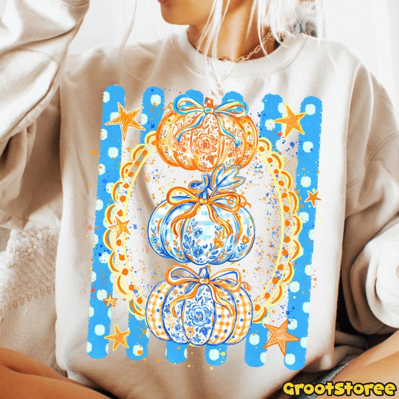 Puede incluir: Una sudadera beige de cuello redondo con un estampado gr&aacute;fico de tres calabazas apiladas decoradas con patrones florales y de cuadros, atadas con lazos naranjas. El dise&ntilde;o incluye un marco circular festoneado, rayas verticales azules y estrellas naranjas.