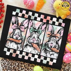 May include: Black decorative pillow featuring three bunny portraits with pink and peach bows, holding carrots. The bunnies are set against a pastel background with an Easter egg design. A checkered border frames the design.