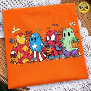 May include: An orange t-shirt featuring a graphic of four characters dressed as ghosts for Halloween. The characters resemble Iron Man, Captain America, Spider-Man, and a classic ghost, each holding a jack-o'-lantern candy bucket.