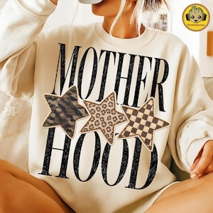 Puede incluir: Una sudadera de cuello redondo color crema con el texto negro MOTHER HOOD en una fuente negrita y desgastada. Tres estrellas están superpuestas en el centro, mostrando un estampado de camuflaje oscuro, un patrón de leopardo marrón y un diseño a cuadros.