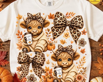 May include: An off-white t-shirt featuring a graphic print of two fluffy highland cows wearing leopard print bows and sweaters. Each cow holds a coffee cup labelled Fueled by Chaos with a pumpkin icon. The design includes autumn leaves and pumpkins.