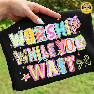 May include: Black towel with the words "WORSHIP WHILE YOU WAIT" in colorful, whimsical lettering. The text features various patterns, including leopard print, plaid, and stripes, with star and bow accents.