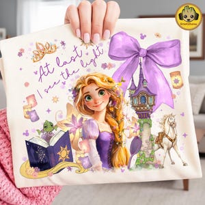 May include: A cream-colored tote bag featuring a watercolor illustration of Rapunzel from the movie Tangled. The design includes a purple bow, a tower, a horse, and the text "At last I see the light."