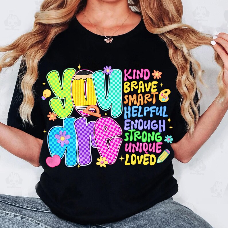 Op de afbeelding: Zwart T-shirt met de woorden "YOU ARE" in kleurrijke, bubbelletters. De woorden "KIND, BRAVE, SMART, HELPFUL, ENOUGH, STRONG, UNIQUE, LOVED" staan verticaal aan de rechterkant.