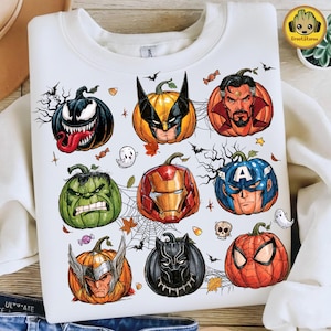 Op de afbeelding: Een wit sweatshirt met ronde hals voorzien van een grafische print van negen pompoenen versierd als superheldenpersonages, waaronder Venom, Wolverine, Doctor Strange, Hulk, Iron Man, Captain America, Thor, Black Panther en Spider-Man met spinnenwebben.
