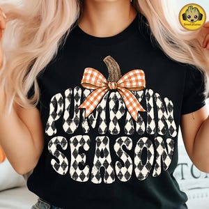 May include: A black short-sleeve t-shirt featuring a large graphic of a pumpkin shape filled with a black and white diamond pattern. An orange and white gingham bow sits at the top of the pumpkin. The text PUMPKIN SEASON is printed in black.