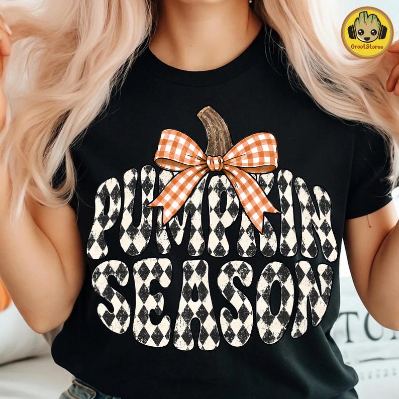 May include: A black short-sleeve t-shirt featuring a large graphic of a pumpkin shape filled with a black and white diamond pattern. An orange and white gingham bow sits at the top of the pumpkin. The text PUMPKIN SEASON is printed in black.