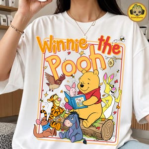 May include: White t-shirt featuring a colorful graphic of Winnie the Pooh and friends reading a book. The design includes the text "Winnie the Pooh" in orange and red, with cartoon characters like Tigger, Piglet, and Eeyore.