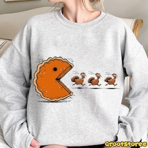 May include: A light gray sweatshirt featuring a humorous Thanksgiving-themed graphic. The design depicts a slice of pumpkin pie chasing three cartoon turkeys. The text "GrootStoree" is at the bottom right.