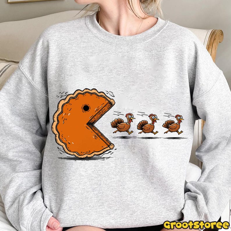 May include: A light gray sweatshirt featuring a humorous Thanksgiving-themed graphic. The design depicts a slice of pumpkin pie chasing three cartoon turkeys. The text "GrootStoree" is at the bottom right.