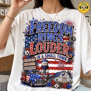 May include: A white t-shirt featuring a graphic print with the text Freedom Rings Louder In A Small Town. The design includes a red, white, and blue truck, a water tower, a barn, fireworks, and wooden signs labelled Faith, Family, and Freedom.