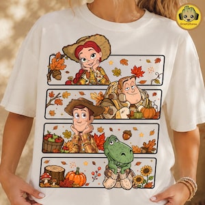 May include: A cream-colored t-shirt featuring a graphic print of four horizontal panels. The panels display cartoon characters Jessie, Buzz Lightyear, Woody, and Rex surrounded by autumn elements like orange leaves, pumpkins, and acorns.
