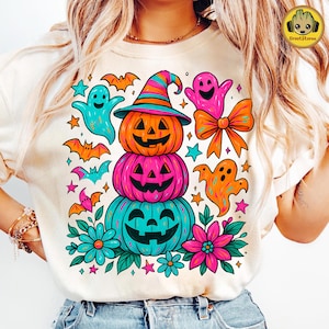 May include: A cream-colored t-shirt featuring a colorful Halloween design. The design includes stacked jack-o'-lanterns, ghosts, bats, stars, and flowers. The top jack-o'-lantern wears a striped witch hat. The design is in bright shades of orange, pink, teal, and blue.