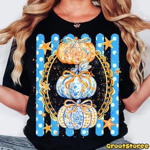 Puede incluir: Una camiseta negra con un gr&aacute;fico de tres calabazas apiladas dentro de un marco ovalado dorado ornamentado. Las calabazas est&aacute;n decoradas con patrones florales y de cuadros en azul y naranja, acentuadas con lazos. Rayas verticales azules y estrellas doradas rodean el marco.