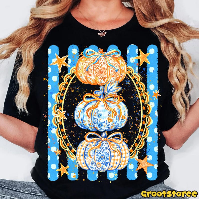 Puede incluir: Una camiseta negra con un gr&aacute;fico de tres calabazas apiladas dentro de un marco ovalado dorado ornamentado. Las calabazas est&aacute;n decoradas con patrones florales y de cuadros en azul y naranja, acentuadas con lazos. Rayas verticales azules y estrellas doradas rodean el marco.