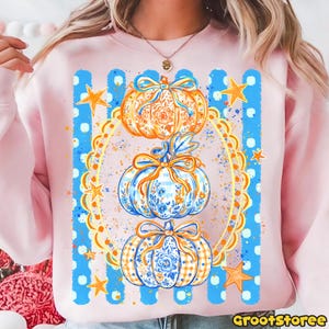 Puede incluir: Una sudadera rosa claro con un estampado gr&aacute;fico de tres calabazas apiladas decoradas con patrones florales azules y naranjas y lazos. El dise&ntilde;o est&aacute; enmarcado por un &oacute;valo festoneado amarillo, rayas verticales azules y estrellas naranjas.