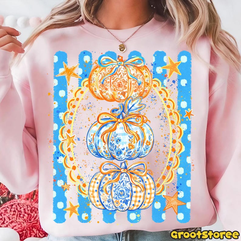Puede incluir: Una sudadera rosa claro con un estampado gr&aacute;fico de tres calabazas apiladas decoradas con patrones florales azules y naranjas y lazos. El dise&ntilde;o est&aacute; enmarcado por un &oacute;valo festoneado amarillo, rayas verticales azules y estrellas naranjas.