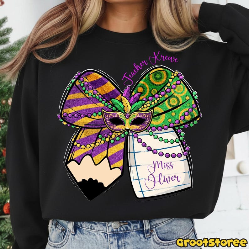May include: Black sweatshirt featuring a large, colorful Mardi Gras bow design. The bow includes purple and gold stripes, green and gold circles, and a decorative mask. The text "Teacher Krewe" and "Miss Oliver" are also visible.