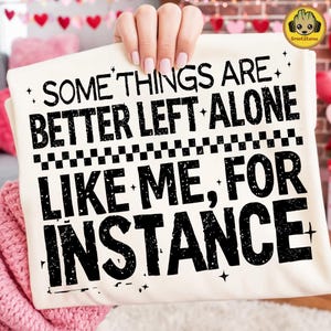 Puede incluir: Una camiseta color crema con el texto "SOME THINGS ARE BETTER LEFT ALONE LIKE ME, FOR INSTANCE" en negro. La camiseta está sujeta por una persona. El logotipo de Groot Storee está en la esquina superior derecha.