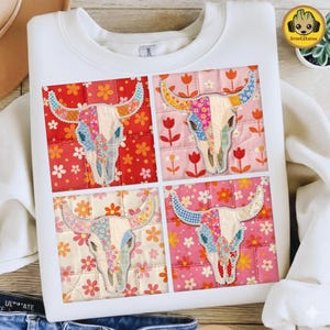 May include: White sweatshirt featuring a patchwork design of four cow skull images. Each skull is decorated with floral and patterned fabric, set against a background of flowers and quilted squares. The design incorporates red, pink, and yellow hues.