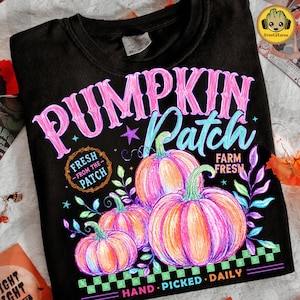 Pode incluir: Uma t-shirt preta com uma estampa gráfica contendo o texto PUMPKIN Patch, FRESH FROM THE PATCH, FARM FRESH e HAND PICKED DAILY. O design inclui três abóboras laranja e rosa, vinhas verdes e uma borda xadrez verde e preta.