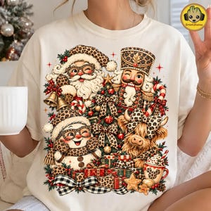 May include: A cream-colored t-shirt featuring a festive graphic of Santa Claus, a nutcracker, a snowman, and a highland cow, all wearing leopard print accessories. The design includes a Christmas tree, wrapped gifts, and bells.