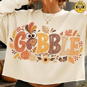 May include: A cream-colored long-sleeve sweatshirt featuring a graphic print with the text GOBBLE. The design incorporates a turkey head, pumpkins, autumn leaves, sunflowers, and acorns in shades of orange, yellow, and brown.