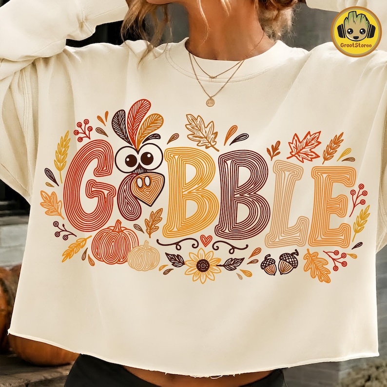May include: A cream-colored long-sleeve sweatshirt featuring a graphic print with the text GOBBLE. The design incorporates a turkey head, pumpkins, autumn leaves, sunflowers, and acorns in shades of orange, yellow, and brown.