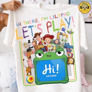 May include: A white t-shirt featuring a colorful graphic of Toy Story characters and a green frog-shaped tablet. The text reads: HI THERE, I'M LILYPAD. LET'S PLAY! and Hi! Let's play! The design includes Woody, Jessie, Buzz Lightyear, and others.